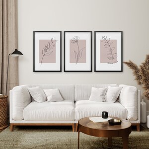 May include: Three black framed prints with a light pink background and black line drawings of leaves and flowers. The prints are hanging on a white wall above a white couch.