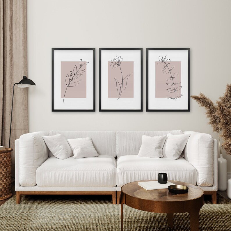May include: Three black framed prints with a light pink background and black line drawings of leaves and flowers. The prints are hanging on a white wall above a white couch.