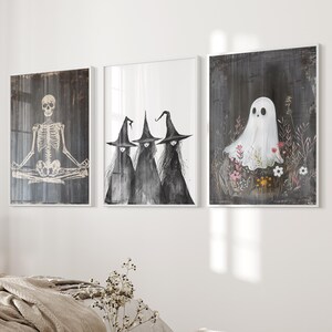 May include: Three black and white witch illustrations with pointed hats and a white ghost illustration with a black outline in a field of flowers. The illustrations are framed in white frames and are hanging on a white wall.