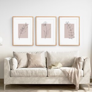 May include: Three framed prints with minimalist line drawings of plants on a blush pink background. The prints are hung on a white wall above a light-colored couch with throw pillows and a blanket.