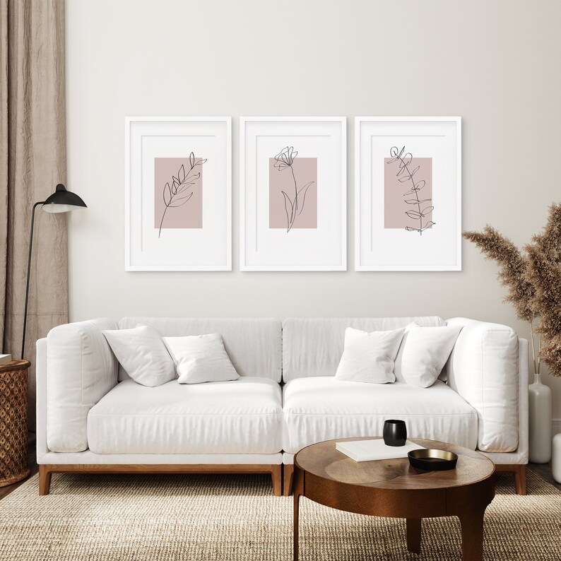 May include: Three framed prints with minimalist line drawings of flowers and leaves on a blush pink background. The prints are hung on a white wall above a white couch.