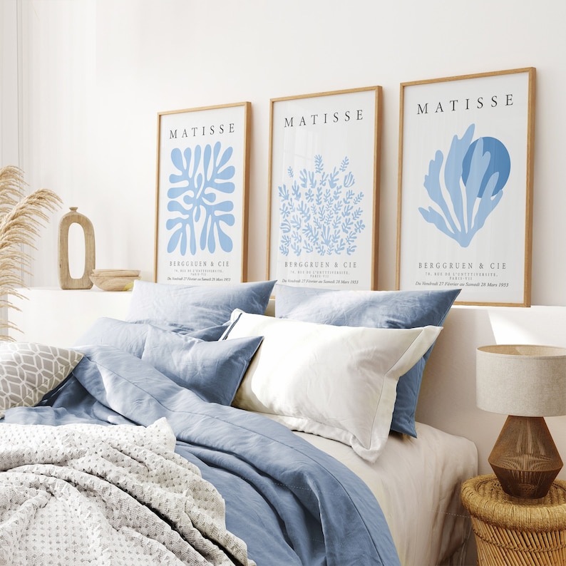 以下が含まれることがあります： 青い抽象的なデザインと白い文字「MATISSE」と「BERGGREEN & CIE」が書かれた額装されたアートプリントが3枚。プリントは白い壁に、青いリネン製のベッドスプレッドの上に飾られています。