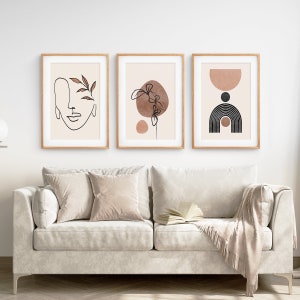 May include: Three minimalist art prints in wooden frames, featuring abstract line drawings and shapes in brown and black on a beige background. The prints are hung on a white wall above a light beige sofa.