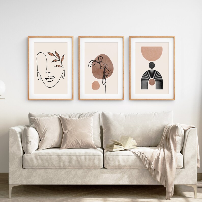 May include: Three minimalist art prints in wooden frames, featuring abstract line drawings and shapes in brown and black on a beige background. The prints are hung on a white wall above a light beige sofa.