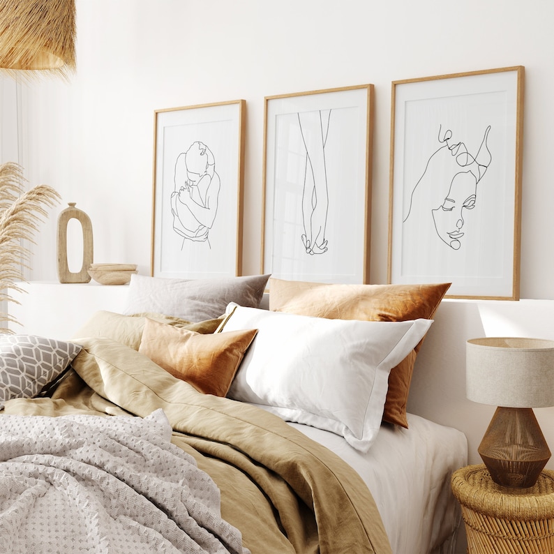 May include: Three framed black and white line drawings of couples in various poses. The drawings are hung on a white wall above a bed with brown linen bedding.