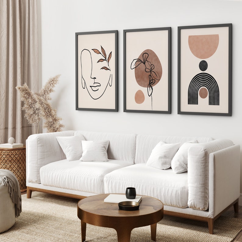 May include: Three framed abstract art prints with minimalist line drawings and muted brown and beige colors. The prints are hung on a white wall above a white sofa.