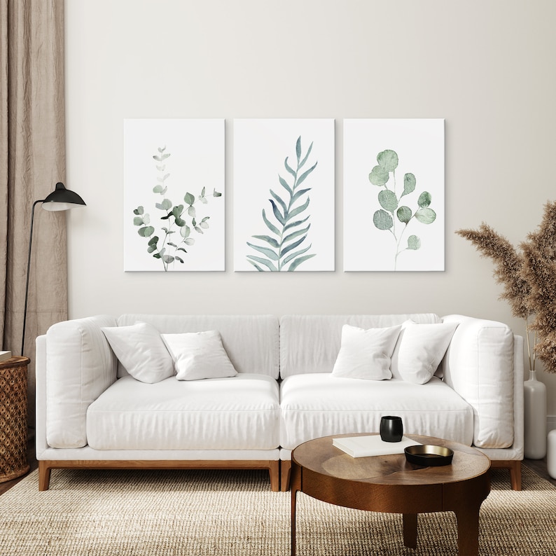 May include: Three watercolor prints of green foliage on white backgrounds. The prints are arranged in a row on a white wall above a white couch.