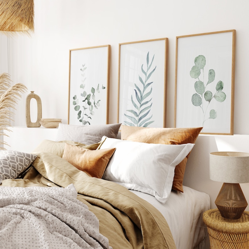 May include: A bedroom with a bed made with white and brown linens, three framed watercolor prints of green leaves on the wall, a wooden side table with a lamp, and a woven rug on the floor.