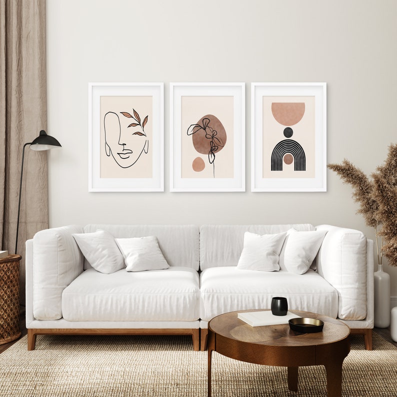 May include: Three minimalist art prints in white frames, featuring abstract line drawings and shapes in shades of brown and black on a cream background. The prints are hung on a white wall above a white couch.
