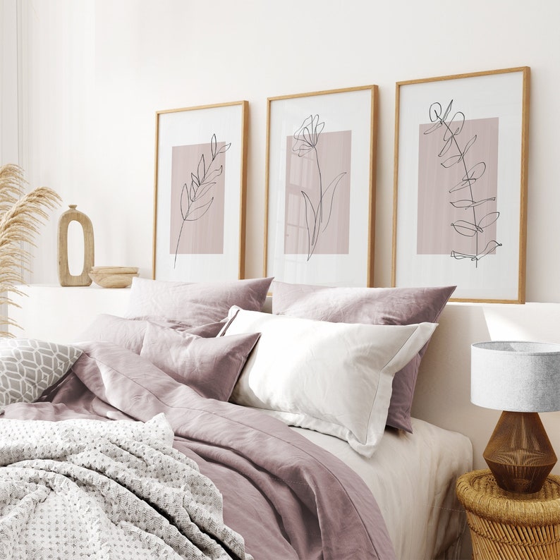 May include: Three minimalist line art prints of floral designs in black on a light pink background. The prints are framed in natural wood and hung on a white wall above a bed with a light pink duvet cover.