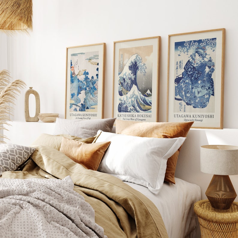 May include: Three framed prints of Japanese woodblock prints. The prints are in blue and white and feature a wave, a mountain scene, and a woman in traditional clothing. The prints are hanging on a white wall above a bed with brown linens.