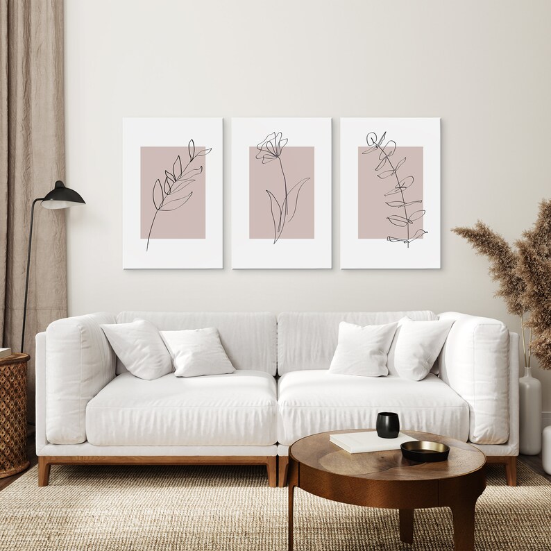 May include: Three minimalist line art prints of floral designs on a blush pink background. The prints are framed in white and hung on a white wall above a white couch.