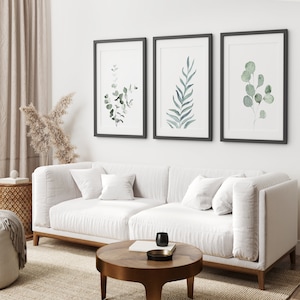 May include: Three framed watercolor prints of green foliage on a white wall above a white sofa with pillows. The sofa is in a living room with a wooden coffee table and a rug.