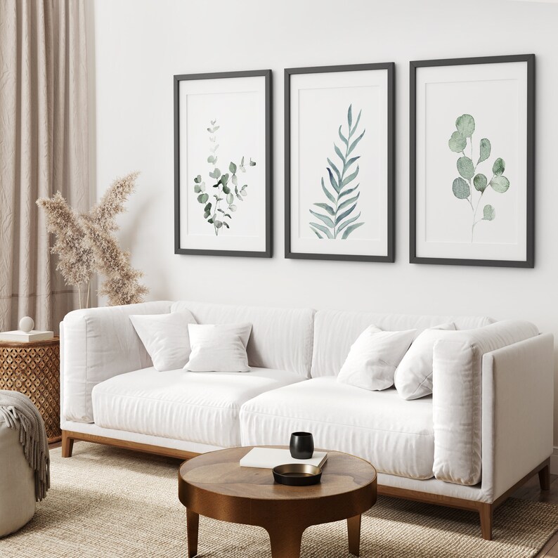 May include: Three framed watercolor prints of green foliage on a white wall above a white sofa with pillows. The sofa is in a living room with a wooden coffee table and a rug.