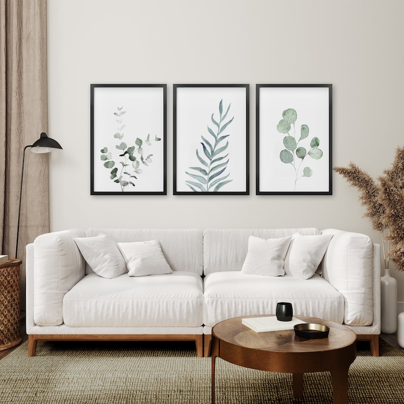 May include: Three framed watercolor prints of green foliage on a white wall above a white sectional sofa with two pillows. A wooden coffee table with a book and a glass sits in front of the sofa.