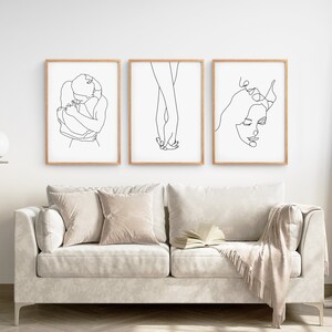 May include: Three black and white line art prints depicting a couple embracing, holding hands, and a woman's face. The prints are framed in natural wood and displayed on a white wall above a white sofa.