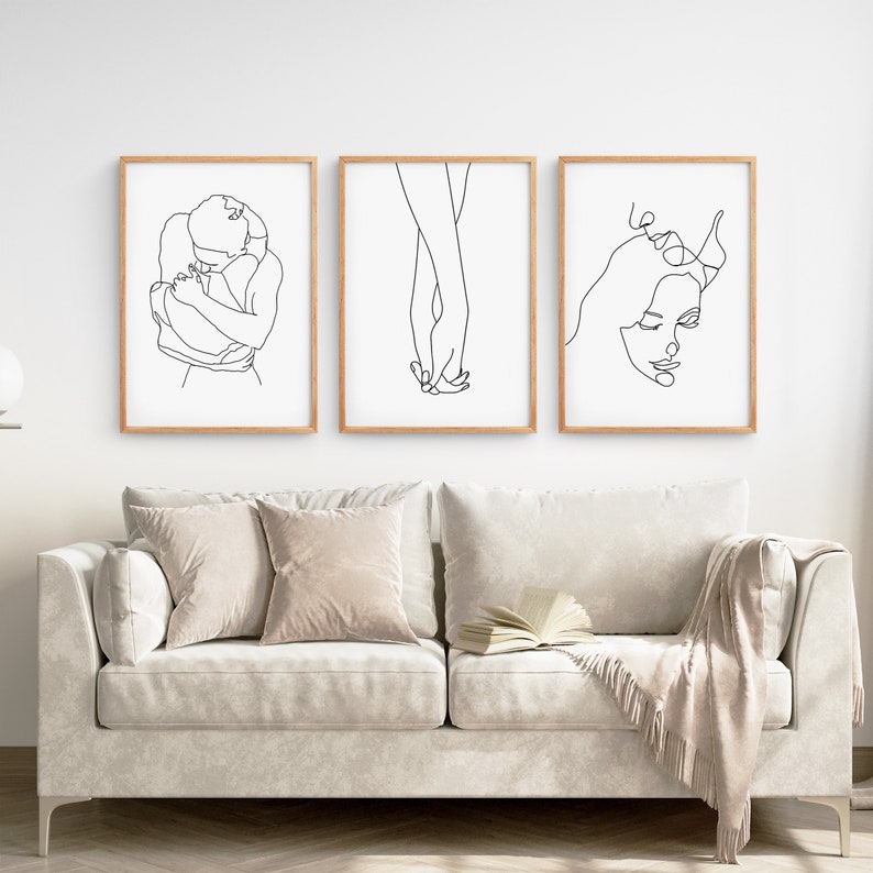 May include: Three black and white line art prints depicting a couple embracing, holding hands, and a woman's face. The prints are framed in natural wood and displayed on a white wall above a white sofa.