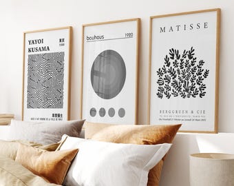 May include: Three framed art prints in wooden frames. The prints display minimalist designs: a black and white pattern, a grey circle with four dots, and a black plant illustration. The prints are displayed above a bed with pillows.