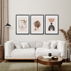 May include: Three framed abstract art prints with a minimalist style. The prints feature line drawings of a face, a flower, and geometric shapes in shades of brown and black on a light beige background.