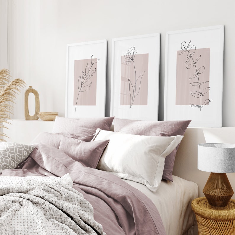 May include: Three framed prints with minimalist line drawings of plants on a blush pink background. The prints are hanging above a bed with a blush pink duvet cover and white pillows.