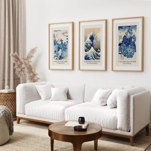 May include: Three framed Japanese woodblock prints, each featuring a different scene. The print on the left depicts a mountain landscape with a blue sky and white clouds. The middle print shows a large wave crashing against a mountain. The print on the right shows a scene with a woman and a cherry blossom tree.