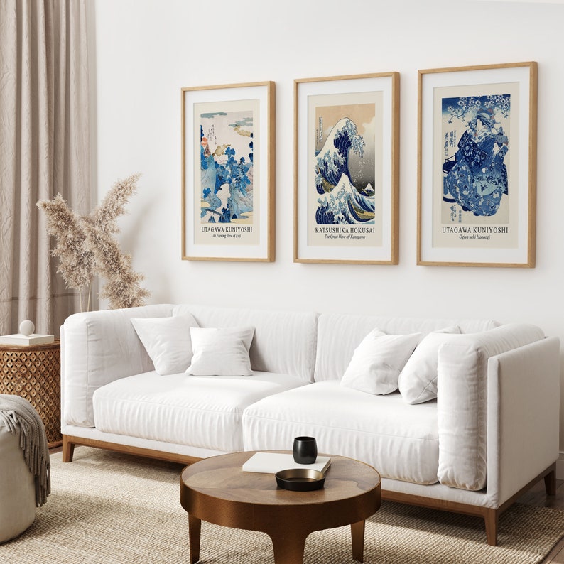 May include: Three framed Japanese woodblock prints, each featuring a different scene. The print on the left depicts a mountain landscape with a blue sky and white clouds. The middle print shows a large wave crashing against a mountain. The print on the right shows a scene with a woman and a cherry blossom tree.