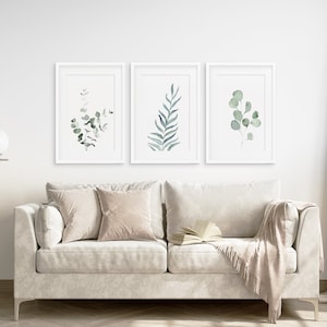 May include: Three framed watercolor prints of green foliage, displayed on a white wall above a light gray sofa with two beige pillows and a beige throw blanket.