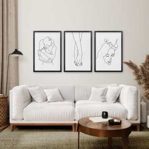 May include: Three black framed line art prints of a couple embracing, holding hands, and a woman's face. The prints are hanging on a white wall above a white sofa.