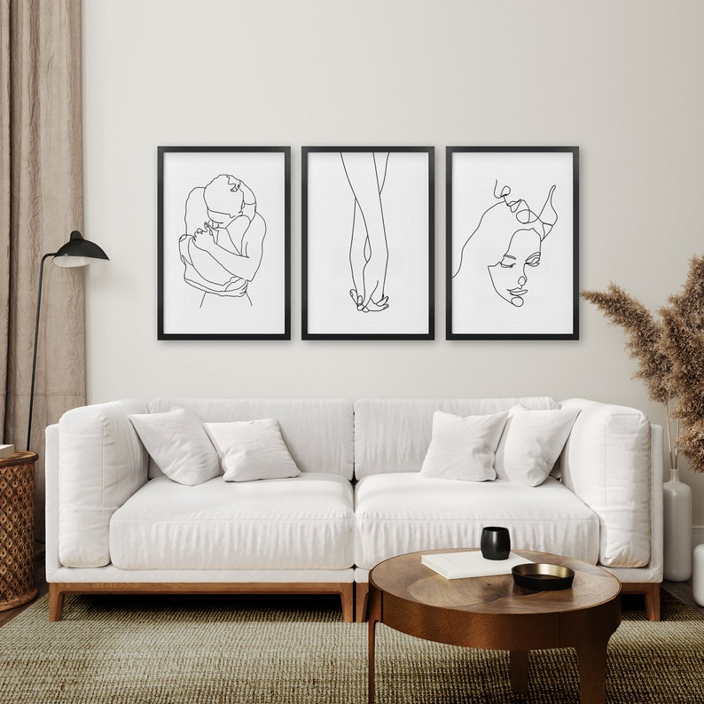 May include: Three black framed line art prints of a couple embracing, holding hands, and a woman's face. The prints are hanging on a white wall above a white sofa.