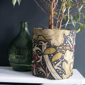 May include: A patterned fabric plant pot in beige, black, and gold, holding a small plant. A green glass olive oil bottle is to the left. The pot's design features a floral pattern. The pot is on a white shelf against a dark blue wall.