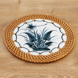 May include: Round coaster with a white ceramic center featuring a blue and white botanical design. The ceramic is surrounded by a woven rattan border. The coaster is designed to protect surfaces from heat and moisture.