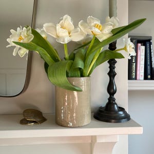 May include: A display of white tulips with green foliage in a beige ceramic vase. A black candlestick with a green candle and a decorative shell sit on a white shelf. A mirror is on the left.