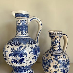 May include: Two ceramic pitchers with a cream-coloured base and blue floral patterns. Both vessels feature a rounded body, a narrow neck, a single handle, and a visible crackle glaze finish throughout the surface of the pottery.