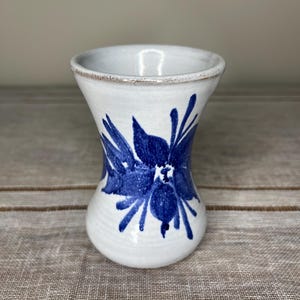 May include: A white ceramic vase with a blue floral design. The vase has a flared top and a rounded body. The blue floral design is painted on the front of the vase. The vase is sitting on a neutral-coloured surface.