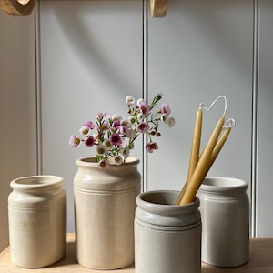 May include: Four antique stoneware jars of varying sizes, arranged on a wooden surface. One jar holds pink and white flowers, another contains several yellow candles. The jars are a creamy beige colour, with a rustic, aged appearance.