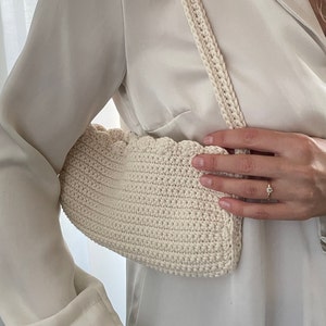 May include: A white crocheted handbag with a scalloped edge and a long strap. The bag is being held by a person wearing a white shirt.