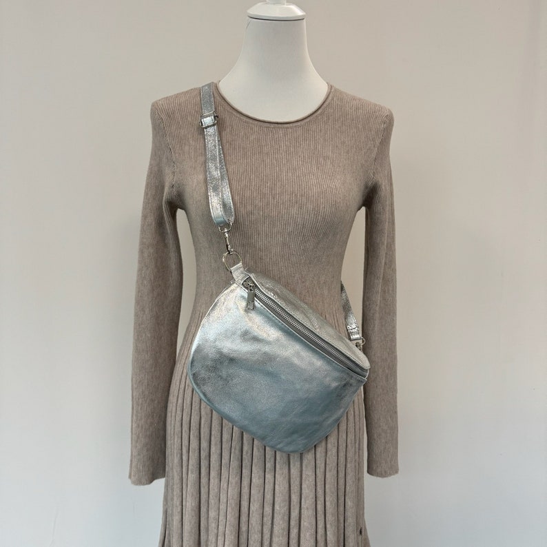 May include: A silver metallic fanny pack with a zipper closure and a long adjustable strap. The bag is worn on a mannequin wearing a beige sweater dress.