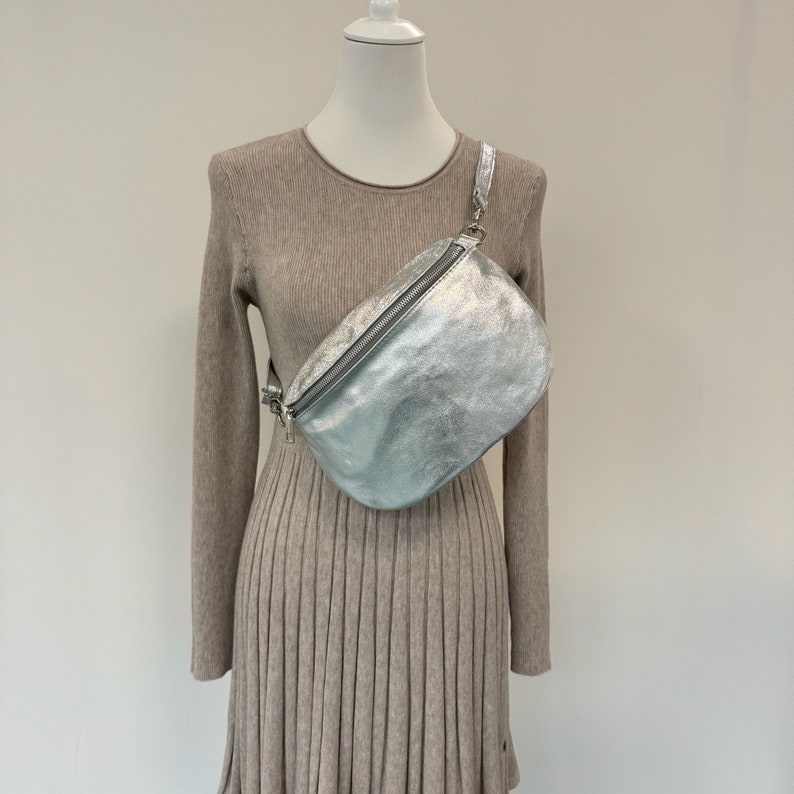 May include: A silver metallic fanny pack with a zipper closure and a silver adjustable strap. The bag is worn on a mannequin wearing a beige sweater dress.