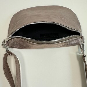 May include: A small, light brown leather crossbody bag with a silver zipper and a silver strap. The bag is open and the inside is lined with black fabric. The label inside the bag reads "GENUINE LEATHER"