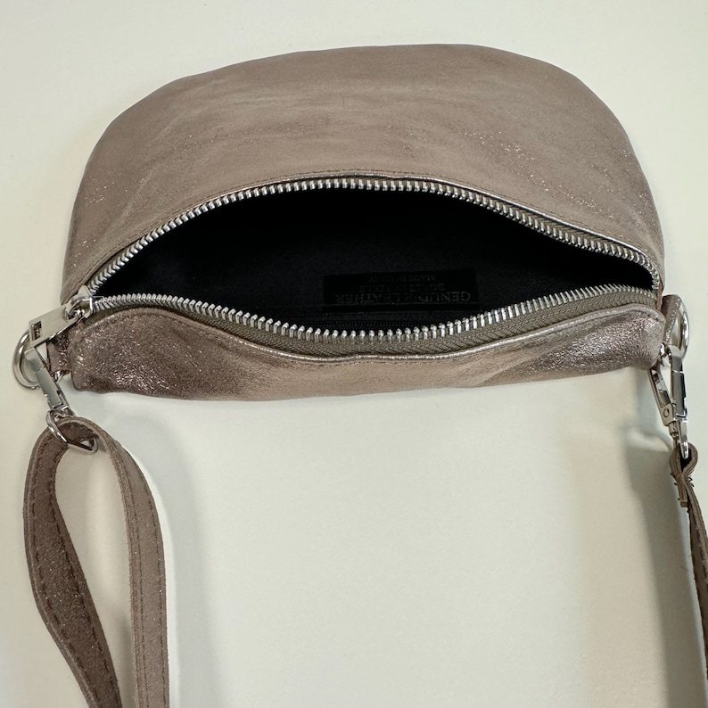 May include: A small, light brown leather crossbody bag with a silver zipper and a silver strap. The bag is open and the inside is lined with black fabric. The label inside the bag reads "GENUINE LEATHER"