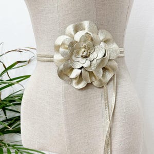Könnte beinhalten: Ein goldfarbener Gürtel mit einer großen, blütenförmigen Schnalle. Der Gürtel ist um eine Schaufensterpuppe gewickelt. Die Blumenschnalle besteht aus geschichteten Blütenblättern. Der Gürtel besteht aus einem dünnen, glänzenden Material.