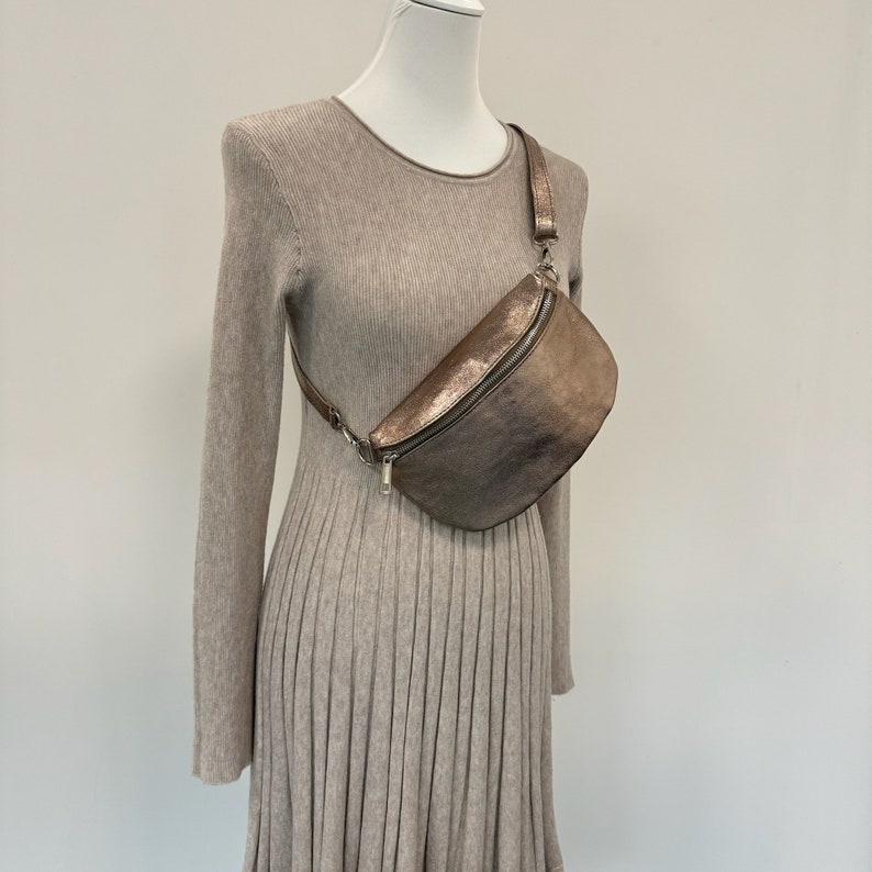 May include: A metallic bronze fanny pack with a silver zipper and strap. The bag is worn on a mannequin wearing a beige sweater dress.