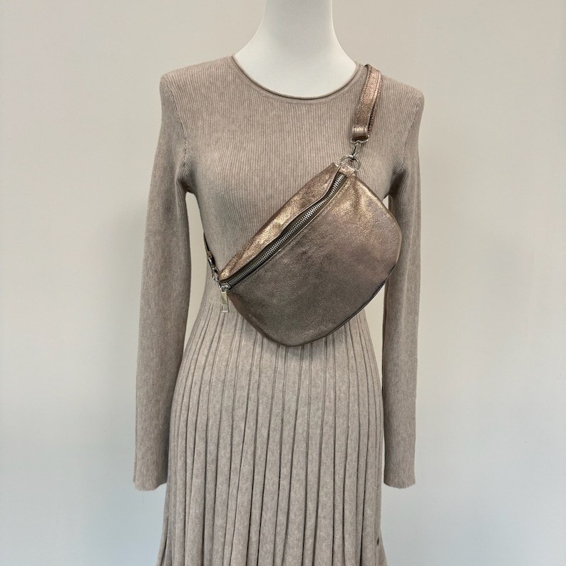May include: A metallic gold fanny pack with a zipper closure and a long adjustable strap. The bag is worn on a mannequin wearing a beige sweater dress.
