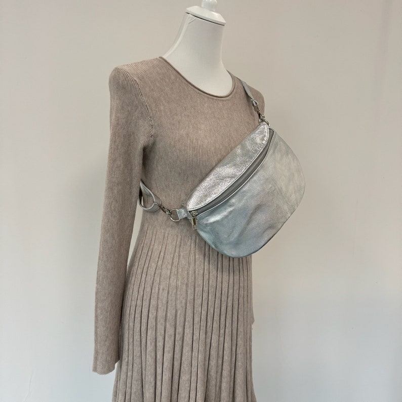 May include: A silver metallic fanny pack with a zipper closure and a chain strap. The bag is worn on a mannequin wearing a beige sweater dress.