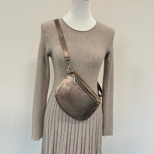 May include: A metallic gold-toned crossbody bag with a zipper closure. The bag is worn over a beige sweater dress.