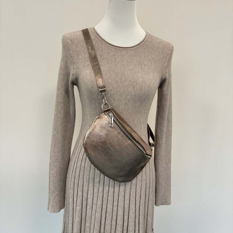 May include: A metallic gold-toned crossbody bag with a zipper closure. The bag is worn over a beige sweater dress.