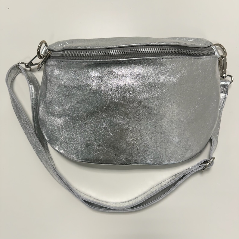 May include: A silver leather fanny pack with a white adjustable strap. The bag has a zipper closure.