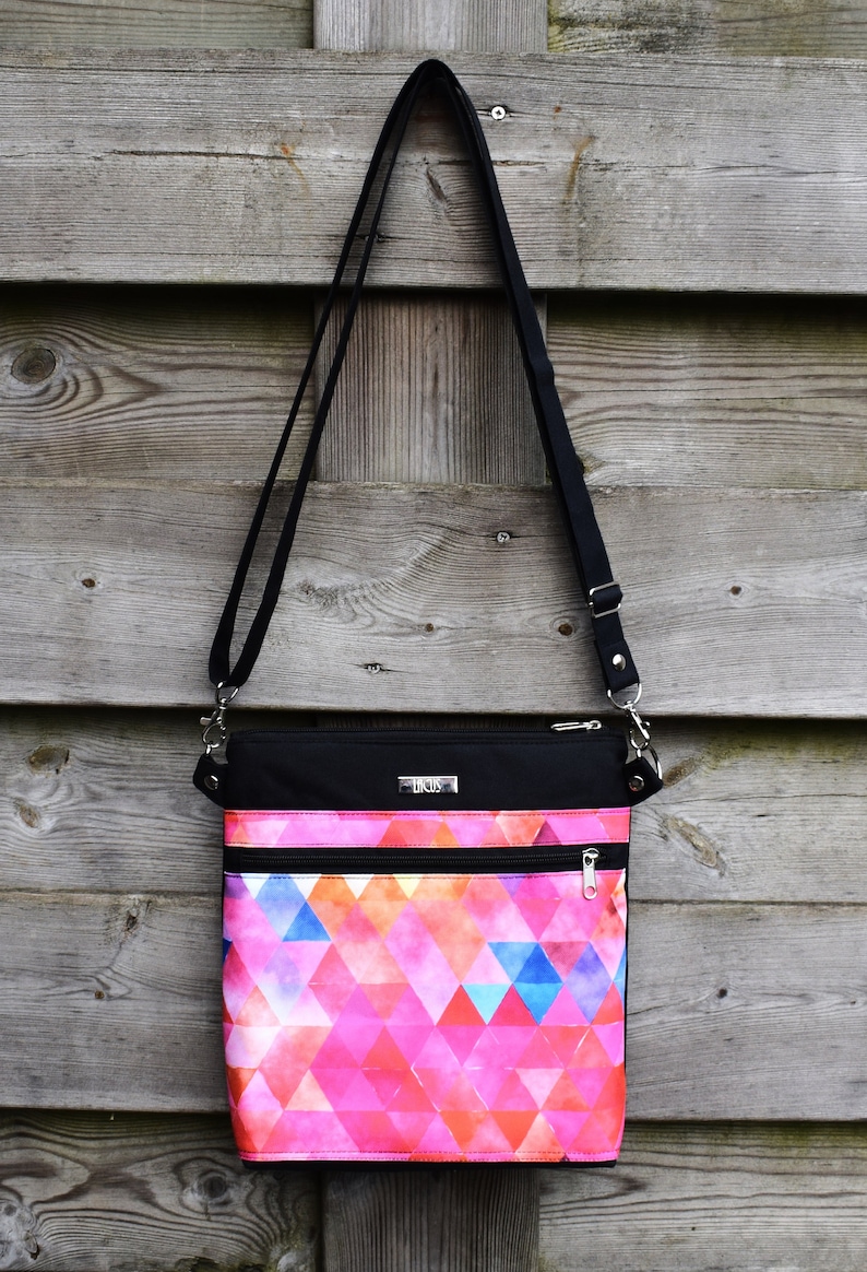 Pu&ograve; includere: Una borsa a tracolla nera con un motivo geometrico rosa e blu. La borsa ha due tasche con cerniera e una tracolla nera.