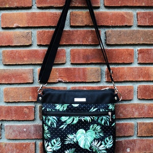 Pu&ograve; includere: Una borsa a tracolla nera con una stampa a foglie tropicali verdi e bianche. La borsa ha una tracolla nera e una chiusura a zip.