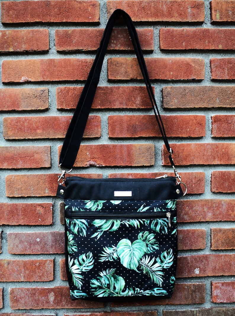 Pu&ograve; includere: Una borsa a tracolla nera con una stampa a foglie tropicali verdi e bianche. La borsa ha una tracolla nera e una chiusura a zip.